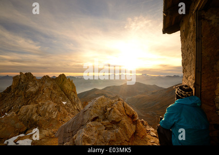Escursionista seduto vicino a capanna e guardando un tramonto mozzafiato dalla cima delle montagne rocciose in italiano - Le Alpi francesi. Foto Stock