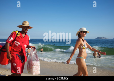 RIO DE JANEIRO, Brasile - 22 gennaio 2014: fornitore di spiaggia camminando accanto alla donna in bikini davanti delle onde sulla spiaggia di Ipanema. Foto Stock