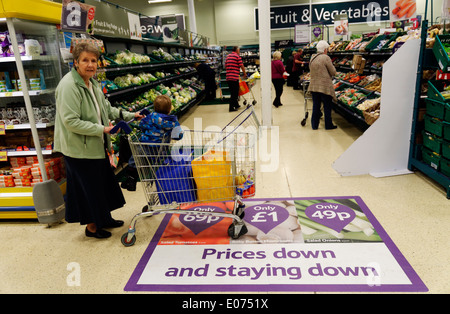 Una vecchia signora shopping nel supermercato Tesco, spingendo un carrello su un pavimento segno annunciando prezzi bassi Foto Stock