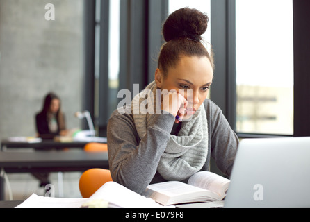 Grave giovane studentessa si concentra sul suo computer portatile mentre studiano in una libreria. Giovane donna seduta a tavola e la lettura. Foto Stock