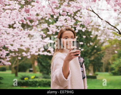 Bella giovane femmina di fotografare con lo smartphone a primavera sbocciano i fiori parco. Femmina caucasica tiro al parco con telefono. Foto Stock