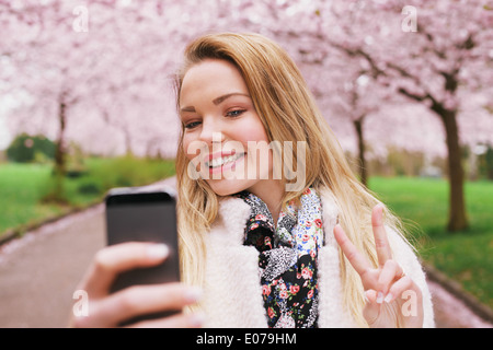 Piuttosto giovane modello femminile gestualità segno di pace mentre prendendo la sua immagine con un telefono cellulare. Bella caucasian giovane donna. Foto Stock