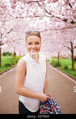 Felice giovane donna che guarda la fotocamera mentre a primavera sbocciano i fiori parco. Modello femminile sul percorso attraverso la fioritura dei ciliegi alberi. Foto Stock