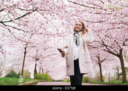 Sorridente giovane donna con telefono mobile per ascoltare musica nel parco. Attraente giovane femmina a molla blossom garden. Foto Stock