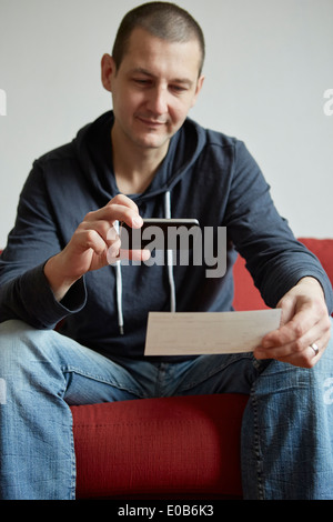 Metà uomo adulto seduto sul divano a fotografare la carta con lo smartphone Foto Stock