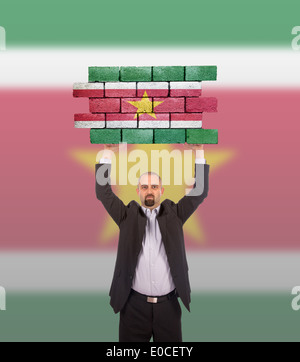 Imprenditore tenendo un grande pezzo di un muro di mattoni bandiera del Suriname isolata sulla bandiera nazionale Foto Stock