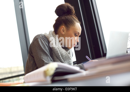 Giovane africano donna americana prendendo appunti da libri per il suo studio. Seduta a tavola con libri e computer portatile per informazioni. Foto Stock