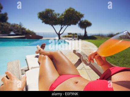 Modello femminile in bikini rosso che giace su una sedia a sdraio a bere un succo di frutta in una giornata di sole da piscina. Giovane donna a prendere il sole. Foto Stock