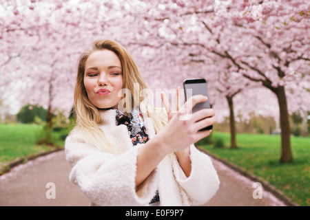 Carino giovane donna gesti segno di pace mentre prendendo la sua foto con il telefono cellulare. Caucasian modello femminile a primavera sbocciano i fiori park Foto Stock