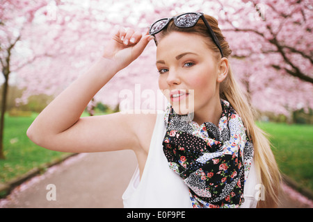 Giovane attraente modello femminile in posa a spring park. Casual giovane donna che indossa gli occhiali da sole e sciarpa guardando la fotocamera. Foto Stock