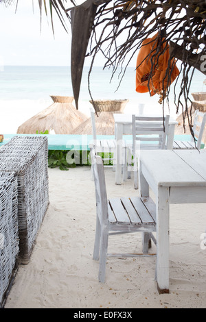 Legno bianco tavoli e sedie sulla superficie sabbiosa con vista oceano e tetti di paglia in background in Tulum, Messico Foto Stock