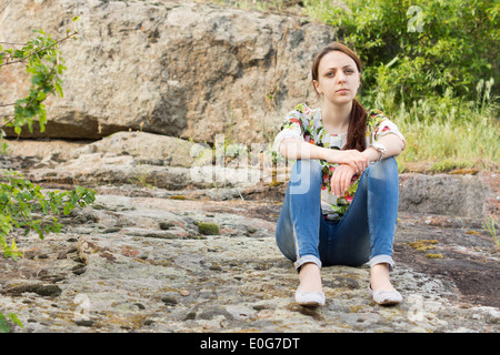 Lonely bella giovane donna con una espressione seria seduta sulle rocce della montagna godendo di un momento di pace e solitudine Foto Stock