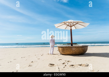 Donna che indossa abiti tipici e cappello di paglia che guarda al mare verso imbarcazioni da pesca, costa di Mui Ne, Sud Vietnam, Vietnam, un Foto Stock