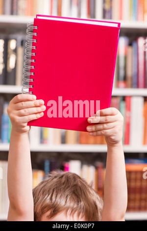 Un giovane bambino azienda libro rosso sopra la testa in una libreria. Foto Stock