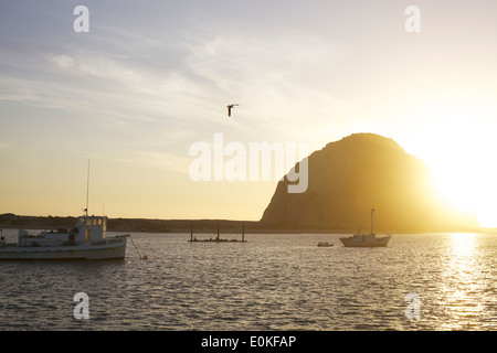 Barche di galleggiare sull'acqua come il sole che tramonta dietro al Morro Bay Rock a Morro Bay, California Foto Stock