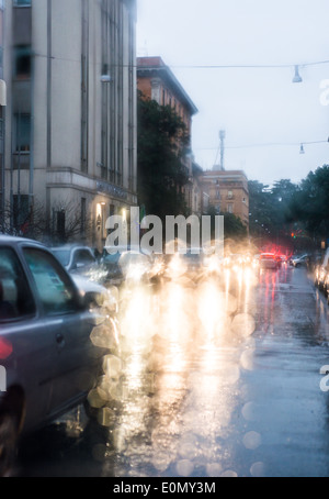 Luce sfocata di vetture visto attraverso un parabrezza bagnato con alcune gocce di pioggia su di esso nella mattina in anticipo di un giorno di pioggia Foto Stock