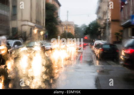 Luce sfocata di vetture visto attraverso un parabrezza bagnato con alcune gocce di pioggia su di esso nella mattina in anticipo di un giorno di pioggia Foto Stock