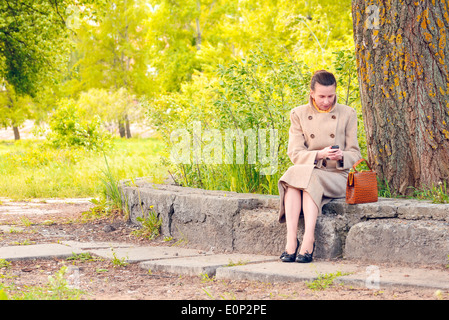 Elegante imprenditrice seduta su un muro di pietra durante una soleggiata giornata di primavera e alla ricerca di un messaggio o la composizione di un numero sul suo telefono cellulare Foto Stock