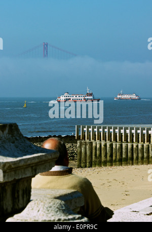 Uomo seduto dal fiume Tago (TEJO) con due traghetti che attraversano e 25 aprile ponte nella nebbia Lisbona Portogallo Europa occidentale Foto Stock