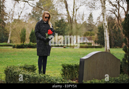 Giovane donna in visita a una persona cara al cimitero pagando rispetta con freschi fiori di rosa. Dolorante femmina al cimitero. Foto Stock