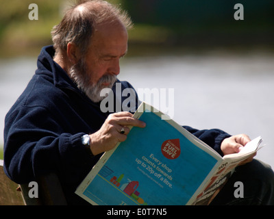 Uomo vecchio seduto su una panchina pubblica lettura di un quotidiano, Cornwall, Regno Unito Foto Stock