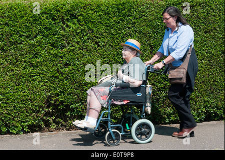 Una signora anziana che indossa un cappello per il sole viene spinto lungo il marciapiede in una sedia a rotelle dal suo accompagnatore o assistente in una giornata di sole. Foto Stock