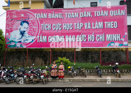 Due fiori donne Hmong in piedi da di un grande tabellone di propaganda dotate di Ho Chi Minh al di fuori di Bac Ha Mercato, Bac Ha, Vietnam Foto Stock