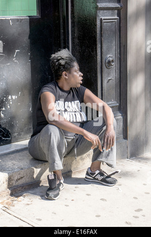 Nero donna indossa suda & sneakers senza lacci si siede sul nono Nona Avenue stoop panhandling in Hells Kitchen Manhattan Foto Stock