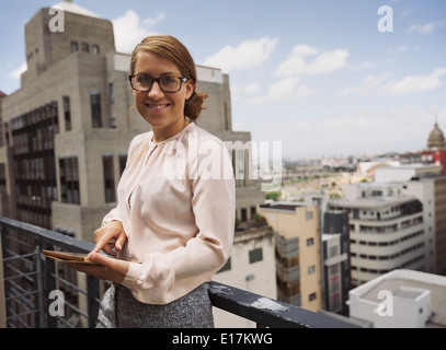 Piuttosto giovane donna in piedi in balcone azienda digitale compressa guardando la telecamera sorridendo. Femmina caucasica in terrazza con vista. Foto Stock
