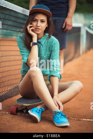 Giovane bella ragazza seduta su uno skateboard sul campo da tennis Foto Stock