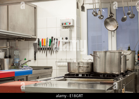 Pulite l'interno di una cucina commerciale con montato a parete utensili e una gamma di differenti pentole in acciaio inox disposti su di una centrale a gas di cottura Foto Stock