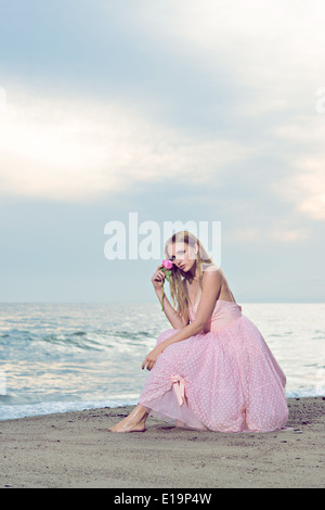 Una giovane e bella donna ritratto seduto su una spiaggia, vestito di una formale rosa 50s abito, tenendo una rosa per il suo volto. Foto Stock