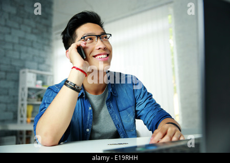 Felice giovane uomo asiatico parlando al telefono al suo posto di lavoro in ufficio Foto Stock