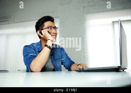 Felice asian uomo seduto al suo posto di lavoro in ufficio e parlare al telefono Foto Stock