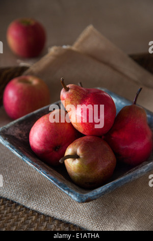 Gruppo di rosso le pere di rustico piatto in ceramica, sulla tela di sacco e vassoio di tessuti, le mele rosse in background, Foto Stock