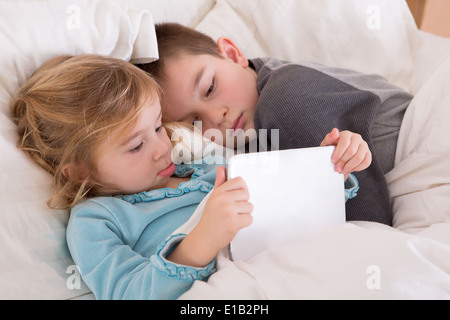 Carino piccolo ragazzo e una ragazza la lettura di una storia prima di addormentarsi in quanto essi si trovano insieme nel letto guardando un computer tablet come essi si preparano ad andare Foto Stock