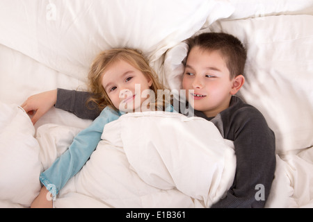 Protezione ragazzo giovane che giace nel letto con la sua sorellina a guardare oltre come lei sta sorridendo fino alla telecamera come si preparano Foto Stock
