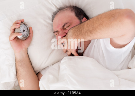 Uomo disteso nel letto a sbadigliare come egli cerca di svegliarsi con la sua sveglia innestata nella sua mano Foto Stock