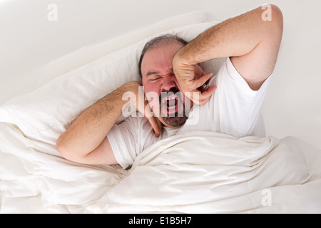 Stanco uomo disteso nel letto di stiramento e sbadigli in uno sforzo per il risveglio come egli dibattiti semplicemente girando su e tornare a dormire in Foto Stock