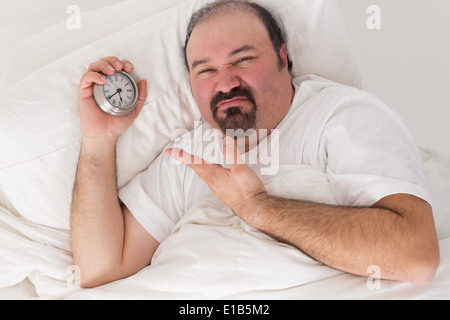 Di mezza età uomo barbuto tenuti svegli da vicini rumorosi che giace nel letto smorfie e indicando il tempo sul suo orologio sveglia Foto Stock