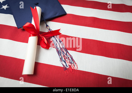 Tappo di graduazione con fiocco e nastro rosso avvolto Diploma in appoggio sulla bandiera americana. Foto Stock