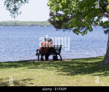 Coppia giovane con le braccia intorno a ogni altro su una panchina nel parco che si affaccia sul lago. Foto Stock