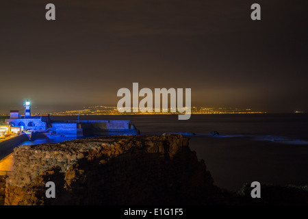 Acre, Israele. Il vecchio porto, il faro e le luci di Haifa in background. Foto Stock