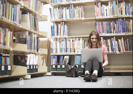 Giovane donna seduta sul pavimento nella libreria utilizzando laptop Foto Stock