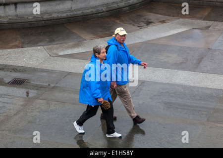 Una coppia di anziani vestiti in blu luminoso impermeabili a piedi attraverso un wet Trafalgar Square, Londra. Foto Stock