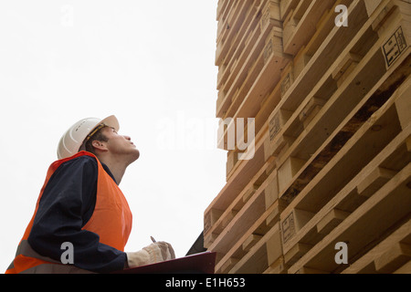 Giovane lavoratore di sesso maschile con appunti cercando fino alla torre di pallet in legno cantiere Foto Stock