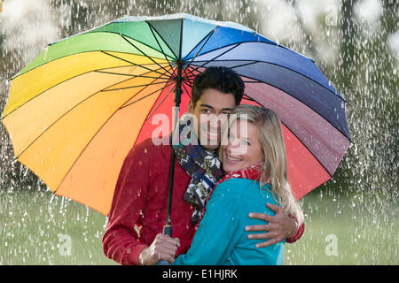 Coppia sorridente sotto un arcobaleno-ombrello colorato godendo la pioggia Foto Stock