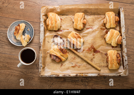Franzbroetchen cotta sulla teglia da forno e la tazza di caffè sul tavolo di legno, vista in elevazione Foto Stock