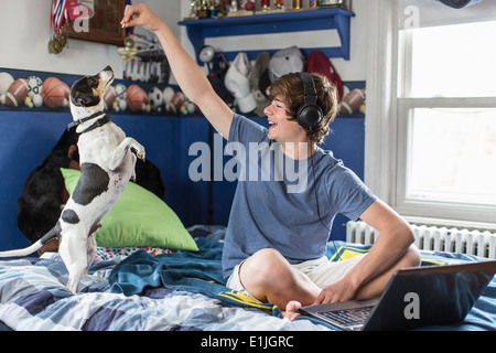 Ragazzo adolescente seduta sul letto con il computer portatile, giocando con il cane Foto Stock