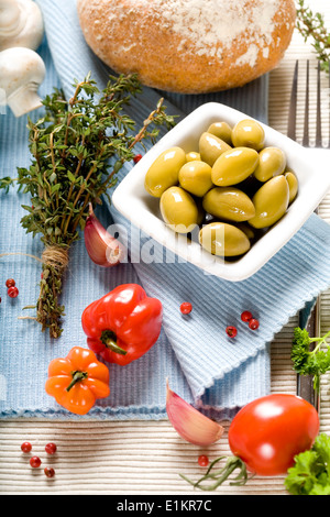 Olive, spezie, pane, verdure e la forcella sul tovagliolo blu con carta bianca Foto Stock